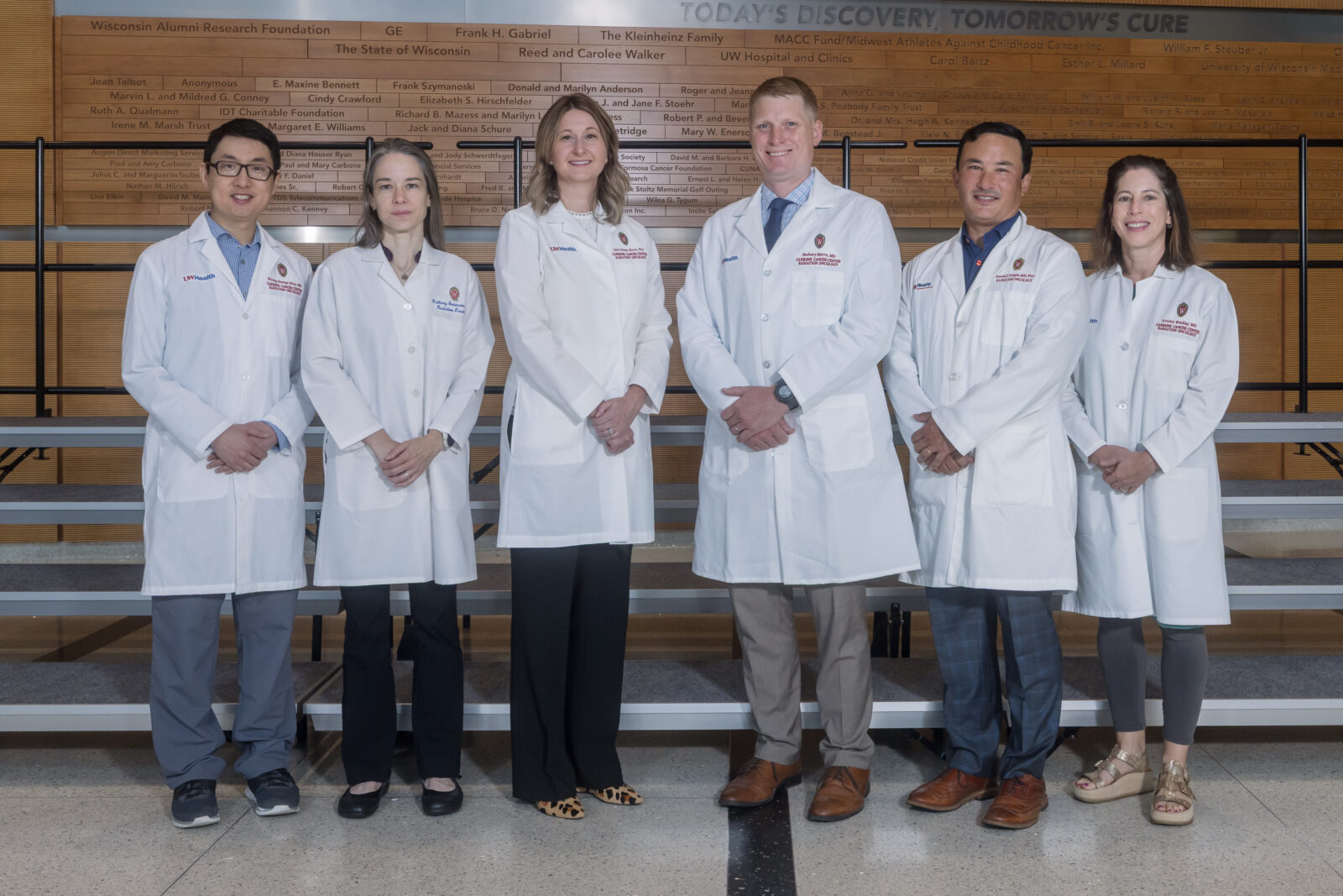 Six doctors in white coats stand in a line with hands folded