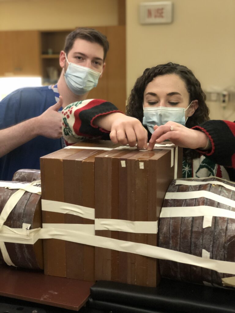 Two physics residents wear masks and work on a wooden phantom