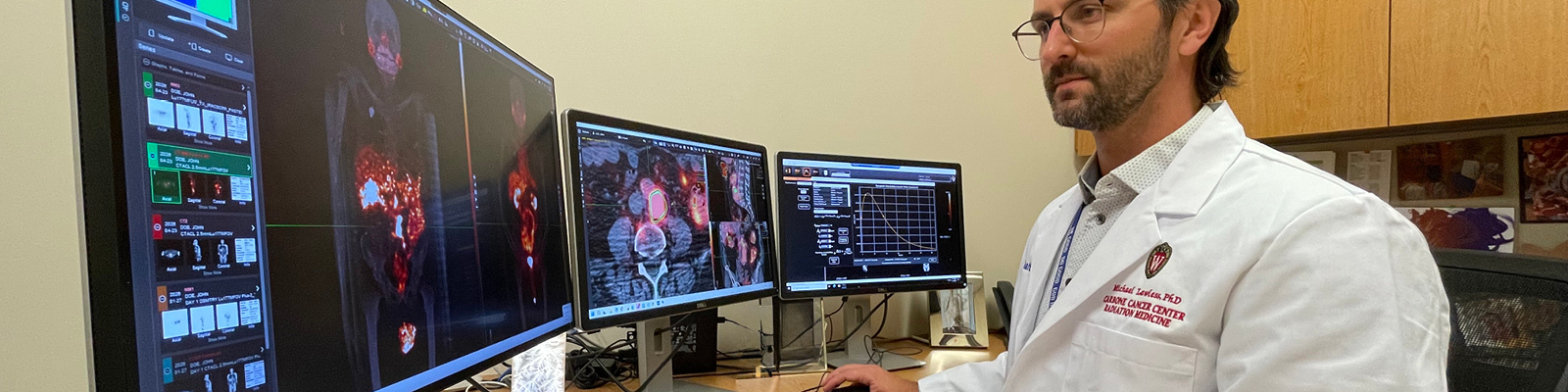 Man in white coat sits at three computer monitors studying scans