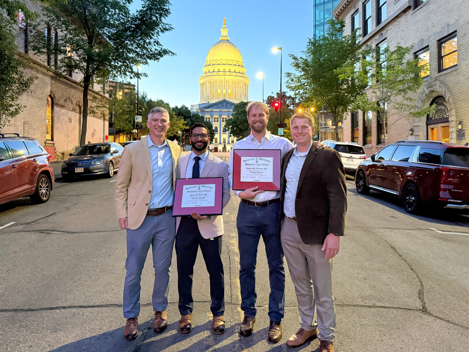 Four men stand in street with capitol behind them. Two graduates hold certificates